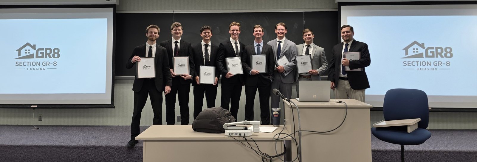 Members of Section Gr-8 Housing in suits holding framed certificates, with the GR8 logo on both projector screens